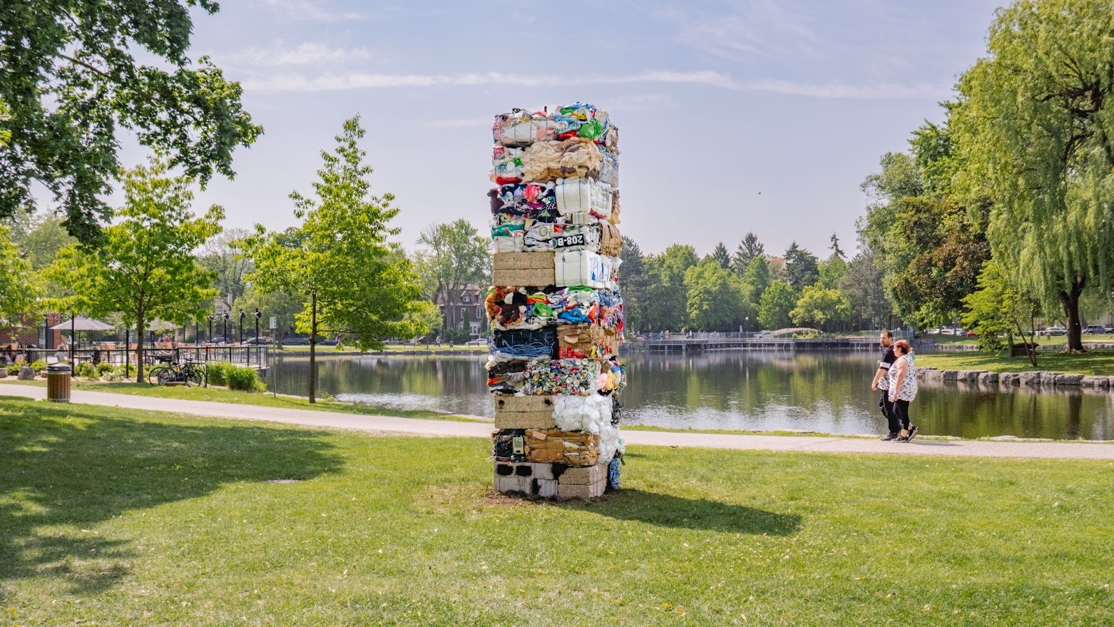 Two people walk on a sidewalk beside a waterway. They are looking toward a tower made from compacted waste for recycling – bottles, fabric, cardboard, and other materials.