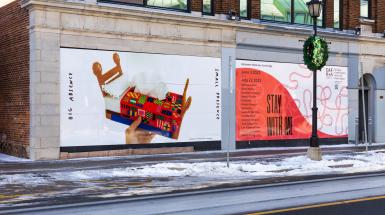 installation view of two large panels on the front of a building featuring an image of a colourful crossed stitched tissue box next to a panel with red biennial branding listing participating artist names