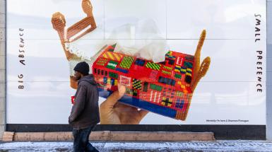 detailed installation view of colourful crossed stiched tissue box shaped like a bed with a person walking in front of it denoting its large scale