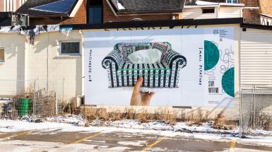 Installation view of primarily green and white crossed stitched tissue box shaped like a couch being held up by a hand. The right border of the image features green biennial branding tied to the Waterloo exhibition zone