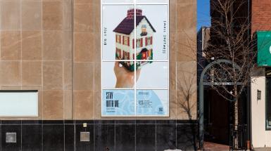 Installation view of a cross stitched tissue box shaped like a house with blue biennial branding at the bottom indicating Cambridge location