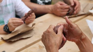 Three sets of hands, sitting around a wooden picnic table working with brown coloured clay