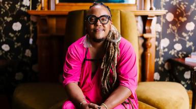 A black person with bold, stylish glasses looks directly at the camera, smiling. He is sitting on a brown chair, wearing a fuschia tunic and one silver hooped earing in his left ear. His long dreaded hair hangs down over his left shoulder. 