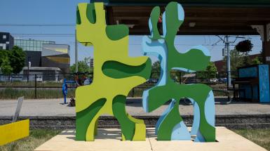 Front View. 8ft sculpture next to visitor's centre, made of overlapping wiggly lime green/forest green shapes fitting like a jigsaw piece into the side of another wiggly forest green/light blue shape.