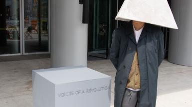 Person standing with pyramid on their head listening to the recording inside. Standing next to a grey plinth that says voices of a revolution.