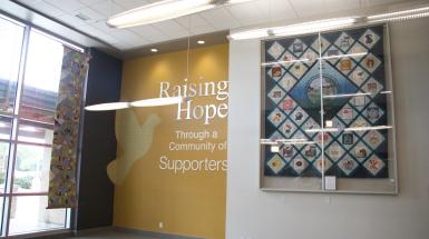 The hanging patchwork quilt is hung in front of a window near another quilt in a box.