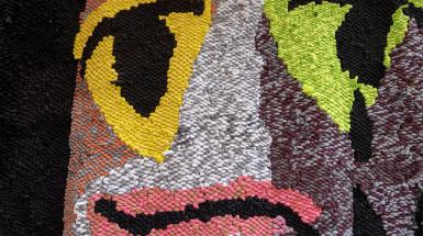 Weaving closeup. Black eyes and lip line with black tears from one eye. Yellow circle around one eye, green circle around the other, pink lips on black and white skin.