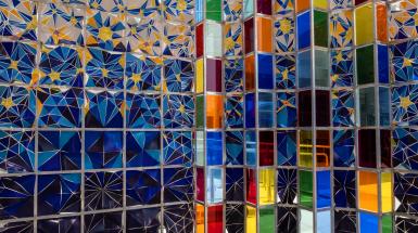 Multicolour mosaic cloth panels in blues and yellows next to many colours of plexiglass panels, all held by an aluminum frame.