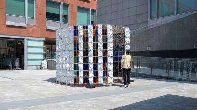 Far shot of the full aluminum structure about 10ft tall, with cloth panels white on the back and coloured plexiglass panes. There is a person standing in front of the structure looking up a it. 