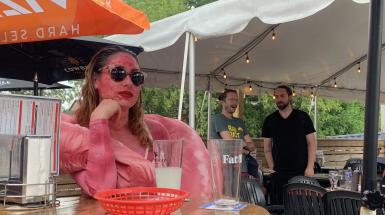 The artist is dressed up as an intestine, wearing sunglasses, a painted pink bodysuit, red platform heels, with a sewn pillow-like intestine scarf/boa. Here the artist is sitting at a restaurant table waiting for food to arrive.