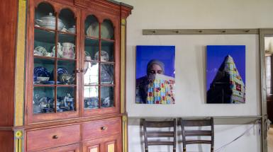 Acrylic panels with blue/indigo background among historical house items, depicting the artist wearing and interacting with their teabag quilt.