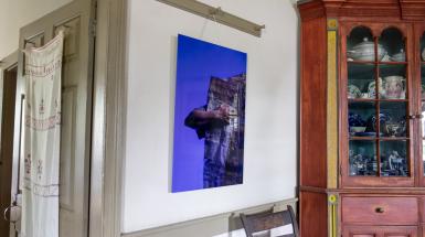Acrylic panels with blue/indigo background among historical house items, depicting the artist wearing and interacting with their teabag quilt.
