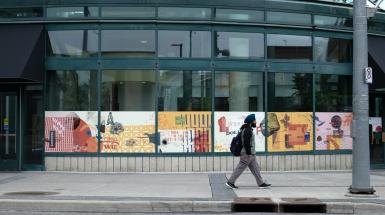 A continuous vinyl collage pasted horizontally across 6 windows, a pedestrian walking by. Collages include pieces of the Kitchener landscape and text on yellow, pink, and cream backgrounds.