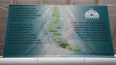 Hanging banner at Kitchener Market focused around a map of the Haldimand Tract with the following text: "The Haudenosaunee have inherited the responsibility to care for the land from time immemorial. Land is a birthright, essential to the expression of our culture. These rights come with specific responsibilities: we can all take from the land what we need to feed and house our families, but we also must assure that the land remains healthy enough to provide for coming generations. Our faith in the Canadian