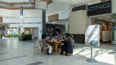 A collection of humanoid figures made from objects sitting around a dinner table in the middle of a mall hallway.