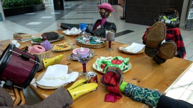 Closer view of the table. Figure on the left is slumped over the table and has a tweed jacket, drum head, basket body and shoes stretchers for hands. Figure at the back seems to be made of cloth with a leather jacket, lantern head, and doll's eyes. Table is full of food made of second-hand objects or odd materials (cloth salad, string spaghetti)