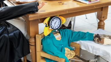 Child-sized figure at a smaller table. It has a clock for a face on a flower head with a blue cardigan and legs made of towel rods put into shoes.