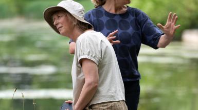 Two of the artists sitting by the water, one speaking to someone out of frame, the other seemingly listening to someone in the other direction.