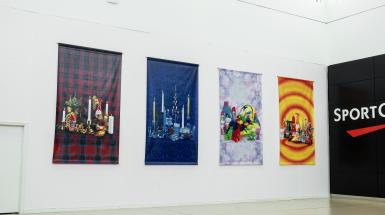 Four large vinyl tapestries hung on a white wall. One is on a red/green checkered background, one on a blue background, one on floral, and one on concentric red/yellow circles. Each tapestry has a collection of similarly coloured objects in the centre.