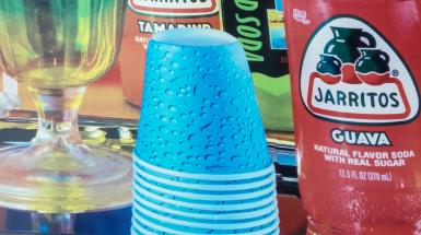 Closeup of bright colourful items arranged in still life - red jarritos bottle, blue dixie cups, green tinted goblet, green soda can, orange jarritos bottle.