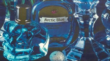 Closeup of darker blue colourful items arranged in still life - a clear bottle of Arctic Blue with blue liquid, a clear perfume bottle with blue liquid, a small speckled white stone/egg, and a clear glass candle holder