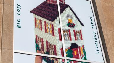 poster of hand holding cross stitched house shaped tissue box cover. And text that reads big loss, small softness.  