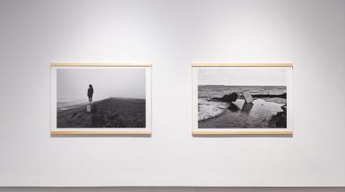 Two photographs hanging on wall. The left image is of a figure standing on a box on the beach, looking to the left to view the water. The image on the right is a figure laying on the beach arms outreached grasping a box as the waves crash into their body. 