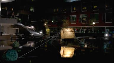 A glowing sculpture of house suspended on water tied to large rocks with rope.