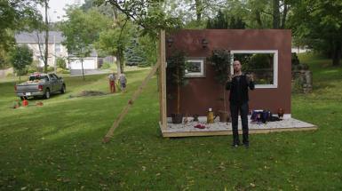A structure made too look like a room with only two walls. Placed in a park. With man speaking in front of it. 