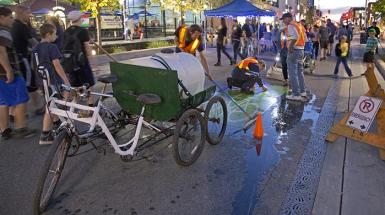 A two seater bike with wagon that holds a water tank. Two men cleaning the bike route sign painted on the ground. 
