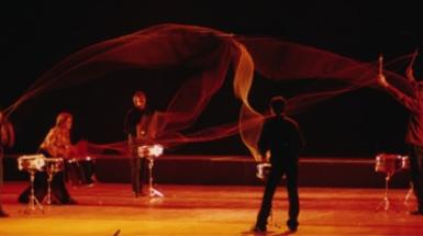 Six individuals playing drums with nylon rope attaching their sticks to other's.