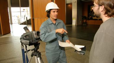 Man in construction hat behind a video camera pointing at a paper talking to another man. 