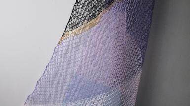 Knitted yarn hung in the shape of a sail boats sail. The yarn is purple, black, white and gold. 