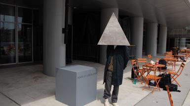 Person in white sneakers, a long navy coat, wearing a grey, textured, triangular helmet that obscures their face, standing beside a grey plith with text that reads "VOICES OF A REVOLUTION"