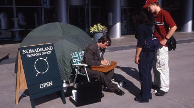 Artist sitting outside tent writing on a paper. Sing to the left says Passports for nomadics. On left two people stand waiting.