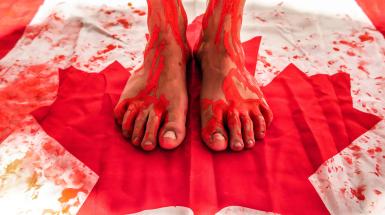 Photo of feet standing on Canadian flag with red paint splattered and dripping