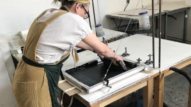 Person in beige overalls standing over a table, pulling black ink through a silkscreen.