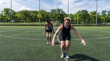 Two women in a sports field with feet together and arms out for balance, hair flying, as though they are jumping with both feet