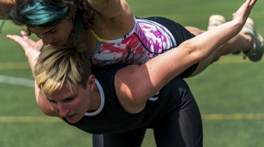 One woman is bent over forward with another woman balanced on her back with arms out and legs extended