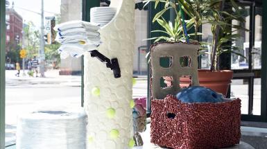 A series of sculptures - one is a box covered in kidney beans holding a bowling ball, another is a white-polka-dot tower filled with stacked plates, with an arm holding towels, and some of the polka-dots are replaced with tennis balls.