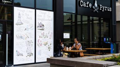 Two people sitting at a table outside cafe pyrus in front of a window vinyl depicting sketches and descriptions of locations or sights in the neighbourhood