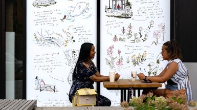 Two people sitting outside cafe pyrus in front of a window vinyl which depicts sketches and descriptions of locations or sights in the neighbourhood
