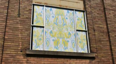 A window in a brick wall-papered over by a yellow and blue pattern on white background. The yellow pattern has images of male and female adult heads, and the overlapping blue has a floating fetus, spread legs, and a floral theme.