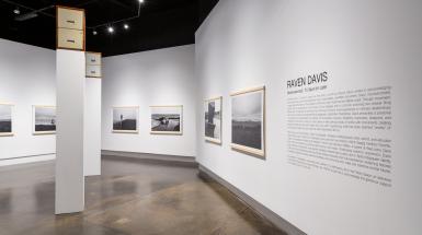 White walls of an art gallery, with black and white photographs framed only on the top and bottom with wood. There are also two double drawer filing cabinets on top of plinths far above head height.
