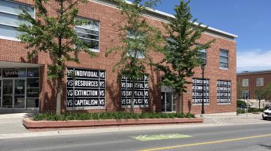 A view of a brick building with windows blacked out, with white text that says in rows "vs individual vs" "vs resources vs" "vs extinction vs" "vs capitalism vs" and many more similar phrases