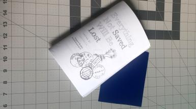 A folded paper booklet sits on a line ruled cutting mat. The cover has the title Everything Not Saved Will Be Lost and several passport stamps