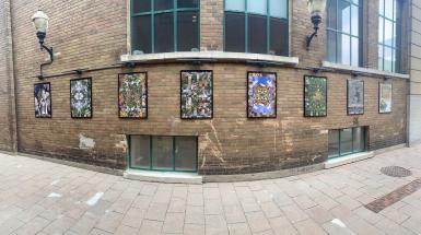 8 digital collage images in a row, all vertical orientation, in black metal frames on the facade of a brick building shot in wide angle