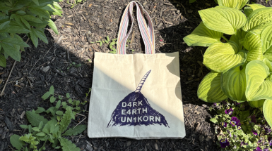 A tote back with a rainbow striped handle lies in a garden bed, surrounded by rocks and flowers. The tote bag, which is natural canvas, shows an indigo screen print of a dirt pile with a unicorn horn poking out. The dirt pile reads "D4RK E4RTH UN1KORN".