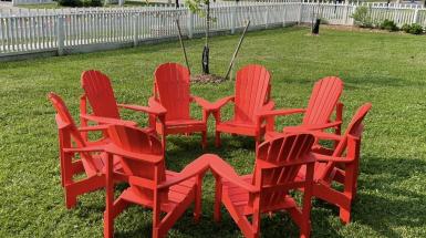 A ring of eight red muskoka chairs are joined at the arms creating an enclosed area, presented on a green turf lawn next to a white picket fence.