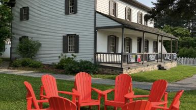 A ring of eight red muskoka chairs are joined at the arms creating an enclosed area, presented on a green turf lawn in front of an older home.